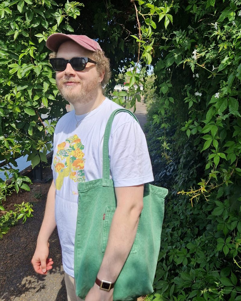 Jack in a pink cap and sunglasses with a green tote bag, by a leafy hedge